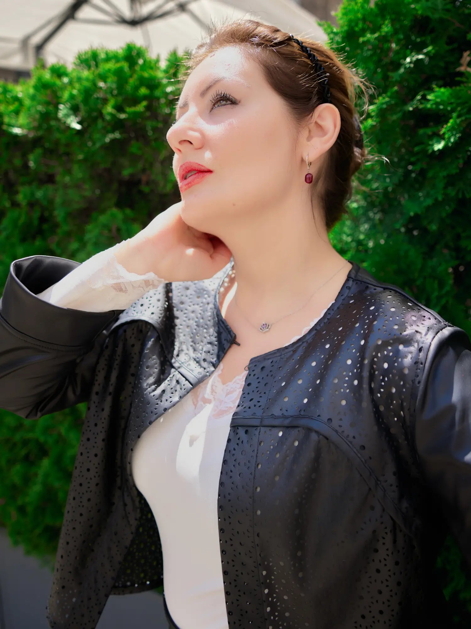 Woman wearing a laser-cut black leather jacket over a white lace-trimmed top, looking upwards with sunlight on her face, standing outdoors in front of greenery