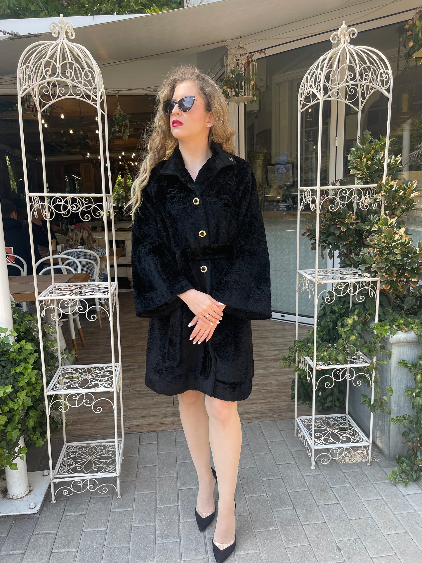 Woman standing confidently in a black textured coat with wide sleeves, gold buttons and a belted waist, styled with black heels and sunglasses in front of a garden café