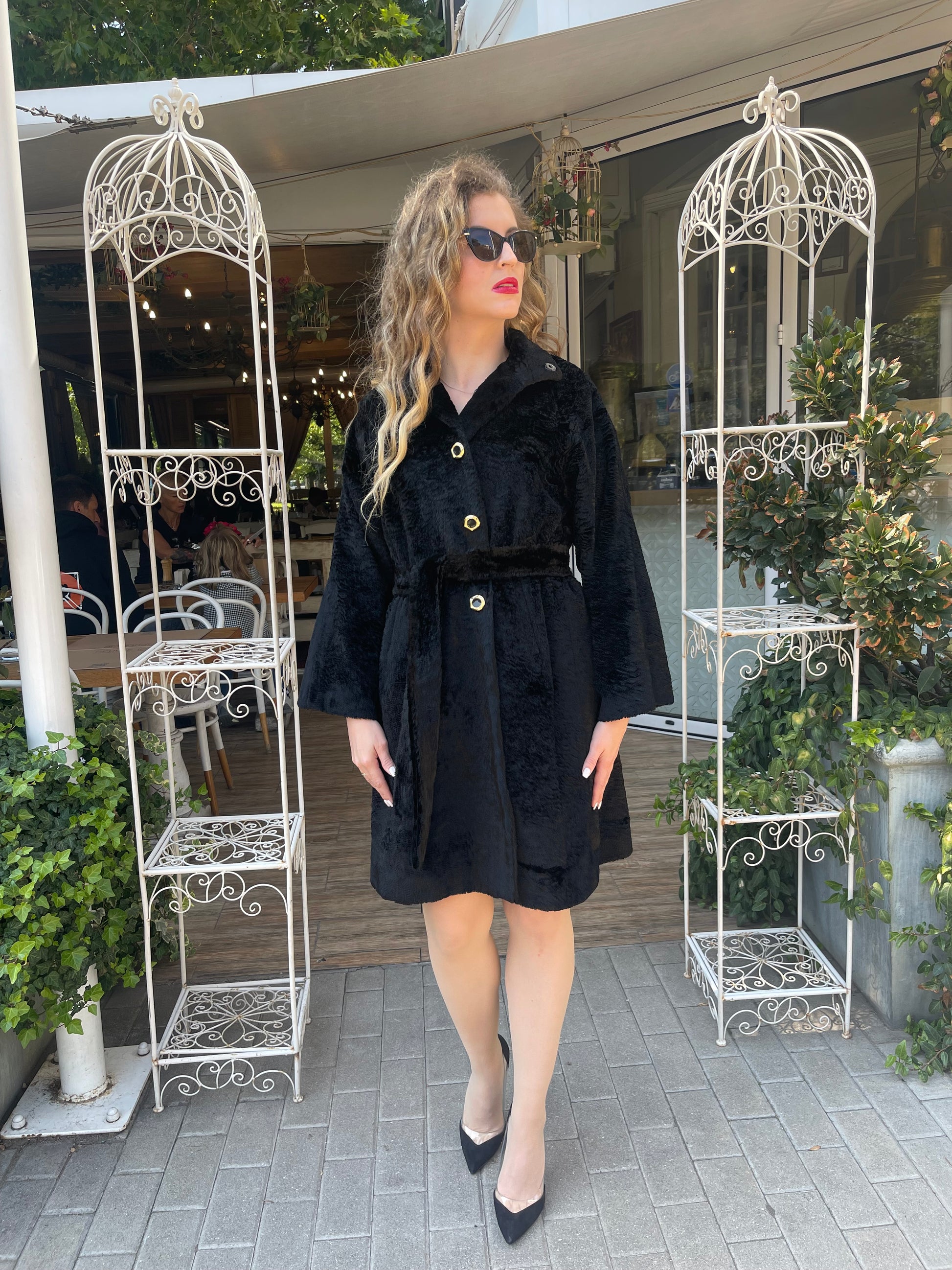 Woman wearing a black textured coat with gold buttons and a belted waist, styled with black heels and sunglasses, standing at the entrance of a stylish outdoor café