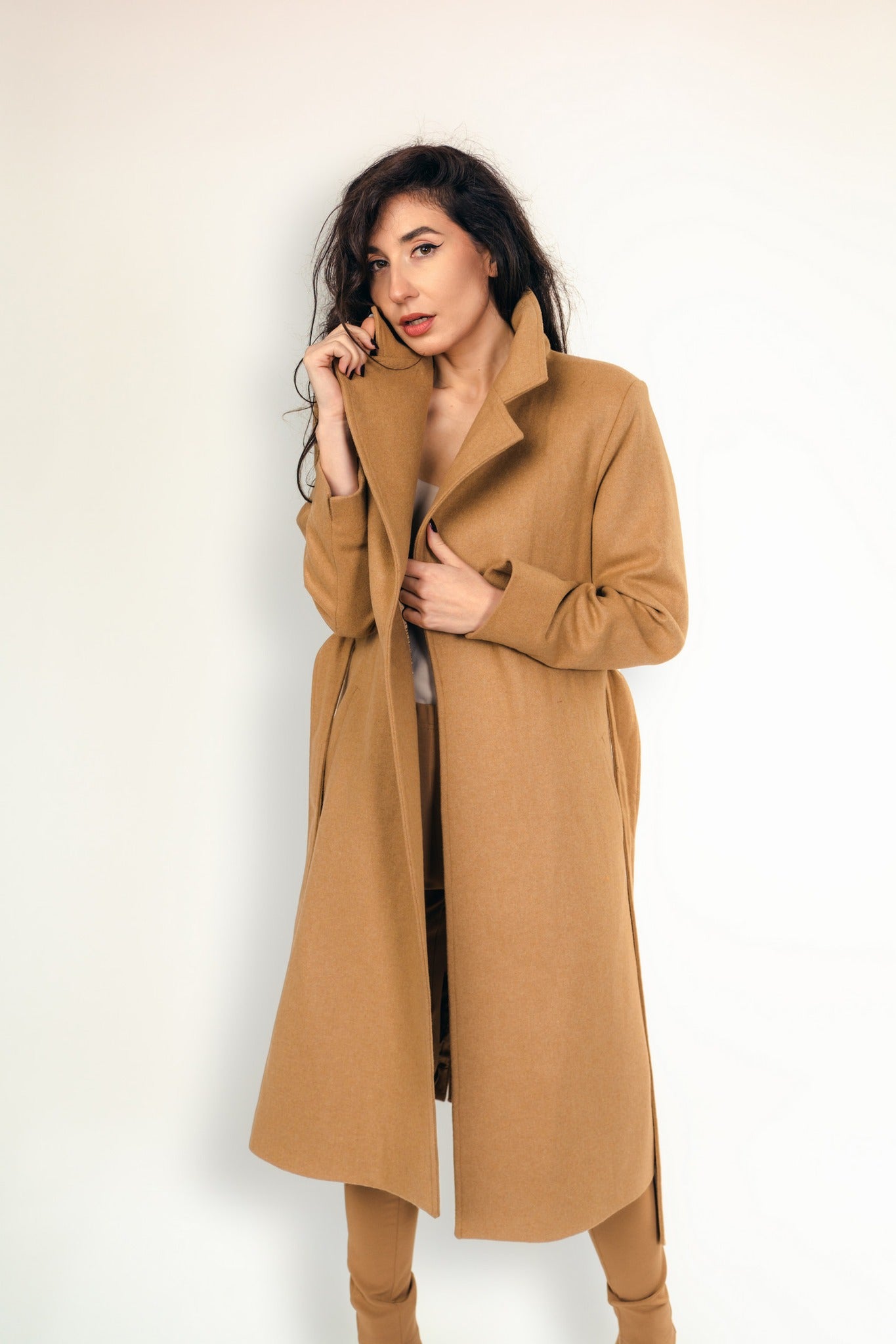 Woman wearing a long camel-coloured wool coat with a structured collar and tailored fit, styled with matching trousers, standing against a neutral background