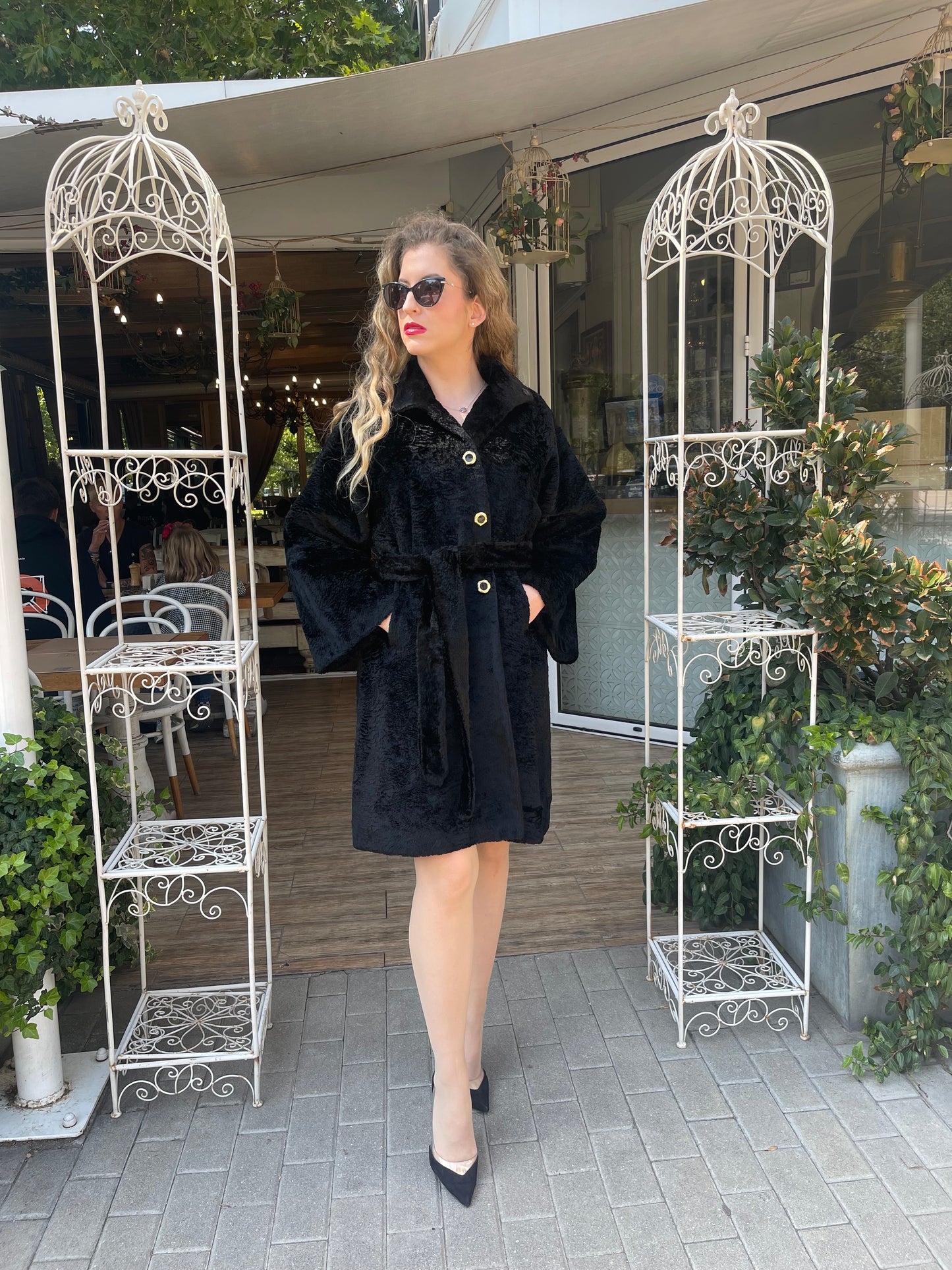 A woman standing in a columned outdoor area wearing a black fur coat with golden buttons, paired with sunglasses and a short-sleeved black top.