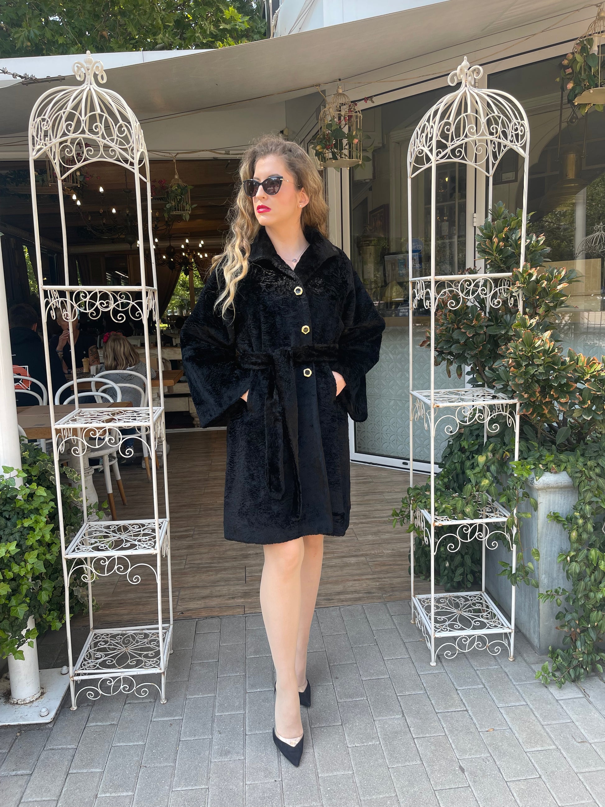 A woman standing in a columned outdoor area wearing a black fur coat with golden buttons, paired with sunglasses and a short-sleeved black top.