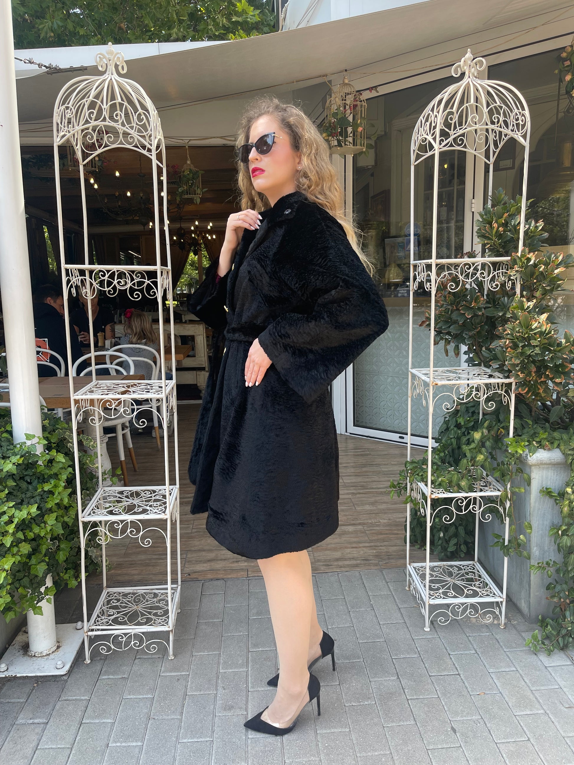 Woman in a black textured mid-length coat with wide sleeves and a belted waist, wearing black heels and sunglasses, standing outside a garden café entrance